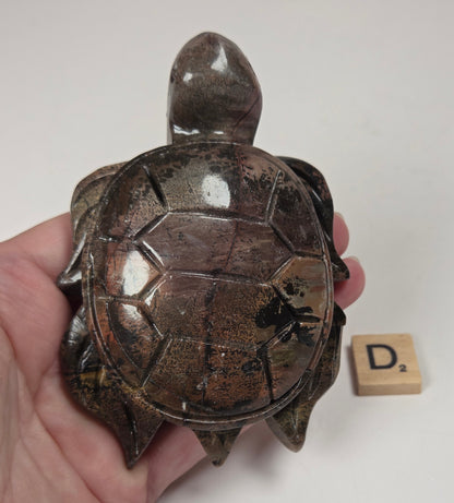 A hand holds a small carved turtle figurine with a glossy, metallic bronze-brown finish. A wooden Scrabble tile showing the letter 'D' is placed next to it on a plain white background.