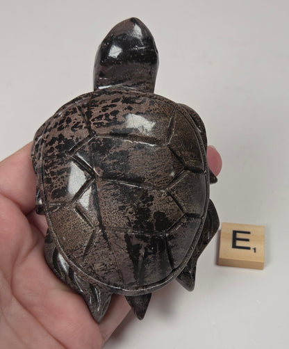 A hand holding a small carved turtle figurine with a glossy dark brown and black shell, shown next to a wooden Scrabble tile with the letter E on a plain light background.