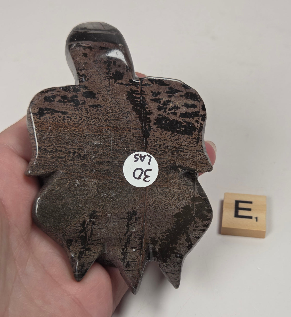 A hand holds a dark brown polished stone or crystal with natural inclusions, shown against a plain light background with a small wooden letter tile beside it.