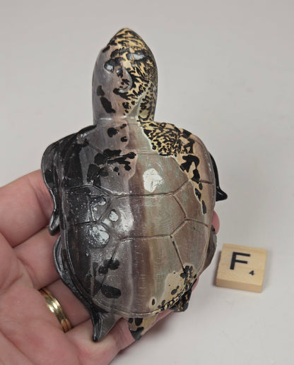 A hand holds a small decorative turtle figurine with a mottled gray, black, and metallic gold shell. A small wooden letter block with the letter 'F' sits beside it on a plain light background.