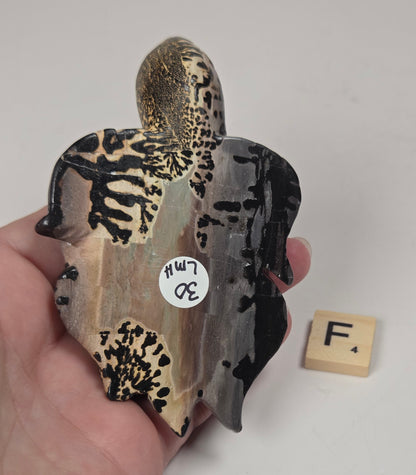 A hand holds a carved stone turtle figurine, showing its natural patterns of black, beige, and gray. A small wooden Scrabble tile with the letter 'F' sits to the right for scale, against a plain off-white background.