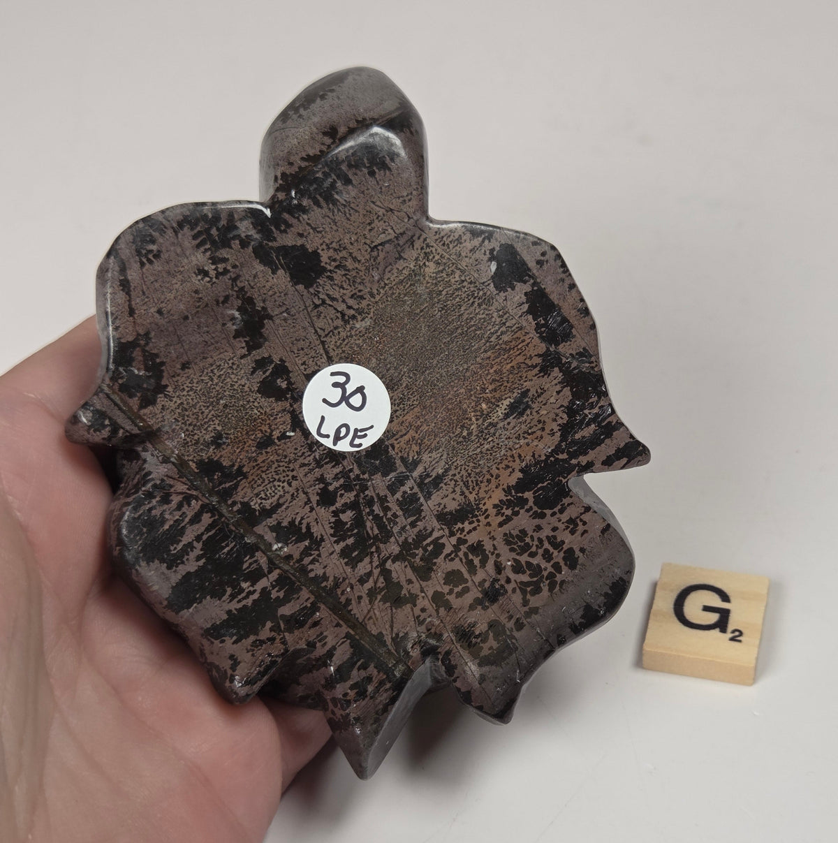 A hand holds a dark, irregularly shaped ceramic object with a mottled brown and black glaze, next to a small wooden letter tile on a plain light background.