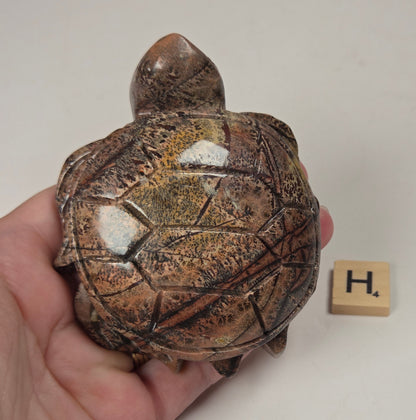 A hand holds a small carved turtle figurine with a glossy brown and gold marbled shell, shown next to a small wooden letter block with the letter 'H' on a plain off-white background.