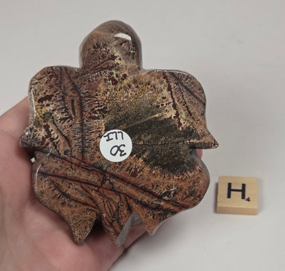 A hand holds a small decorative object shaped like a leaf or shell in a mottled brown and bronze metallic finish, with a small white inventory sticker on it. A wooden Scrabble tile with the letter H sits nearby on a plain light background.