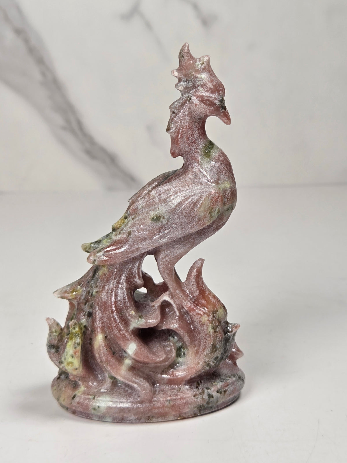 A carved decorative figurine of a rooster standing on flames, made from pinkish stone with green and gray marbling, set against a light marble-patterned background.