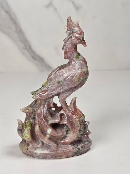 A carved decorative figurine of a rooster standing on flames, made from pinkish stone with green and gray marbling, set against a light marble-patterned background.