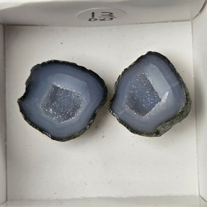 A pair of geode slices with grey outer layers and white crystal-like centers, displayed inside a small white box held by a hand.