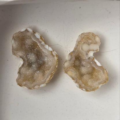 A pair of geode slices with white crystal-like centers, displayed inside a small white box held by a hand.