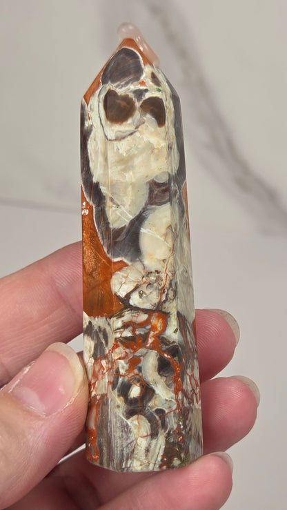 Hand holding a tall, narrow decorative crystal tower made of natural copper pyrite and silver quartz with brown and orange mineral inclusions, standing upright on a white surface against a light gray marble-patterned background.