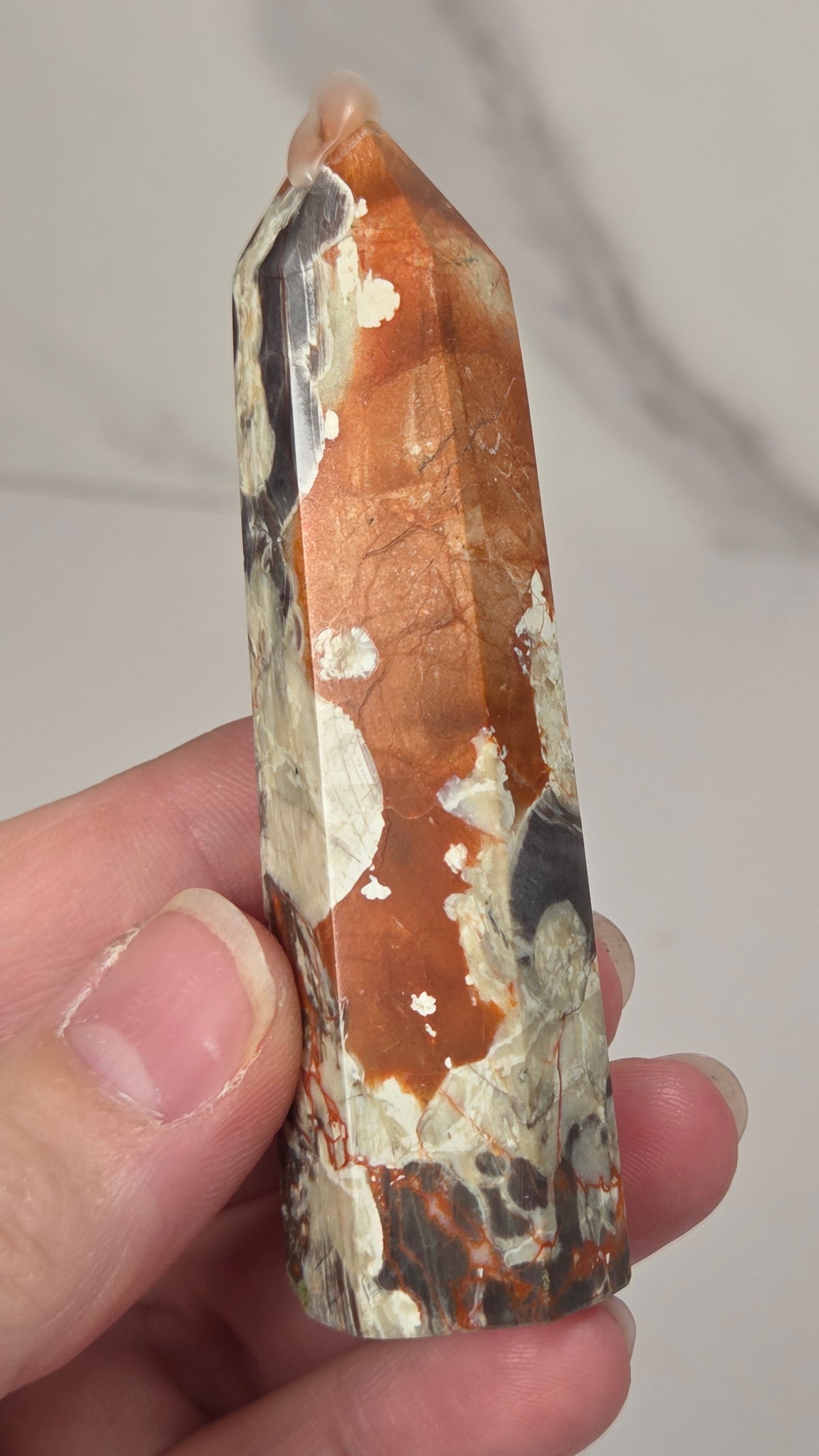 Hand holding a tall, narrow decorative crystal tower made of natural copper pyrite and silver quartz with brown and orange mineral inclusions, standing upright on a white surface against a light gray marble-patterned background.