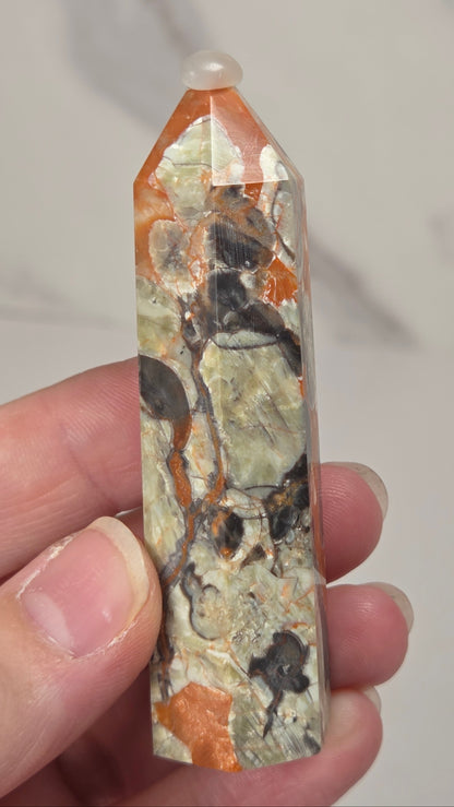 Hand holding a tall, narrow decorative crystal tower made of natural copper pyrite and silver quartz with brown and orange mineral inclusions, standing upright on a white surface against a light gray marble-patterned background.