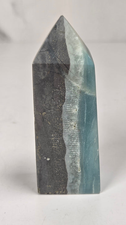 A polished mineral crystal tower with a split dual-color design, featuring light blue and white on one side and dark gray with subtle gold veining on the other, set against a neutral light gray background.
