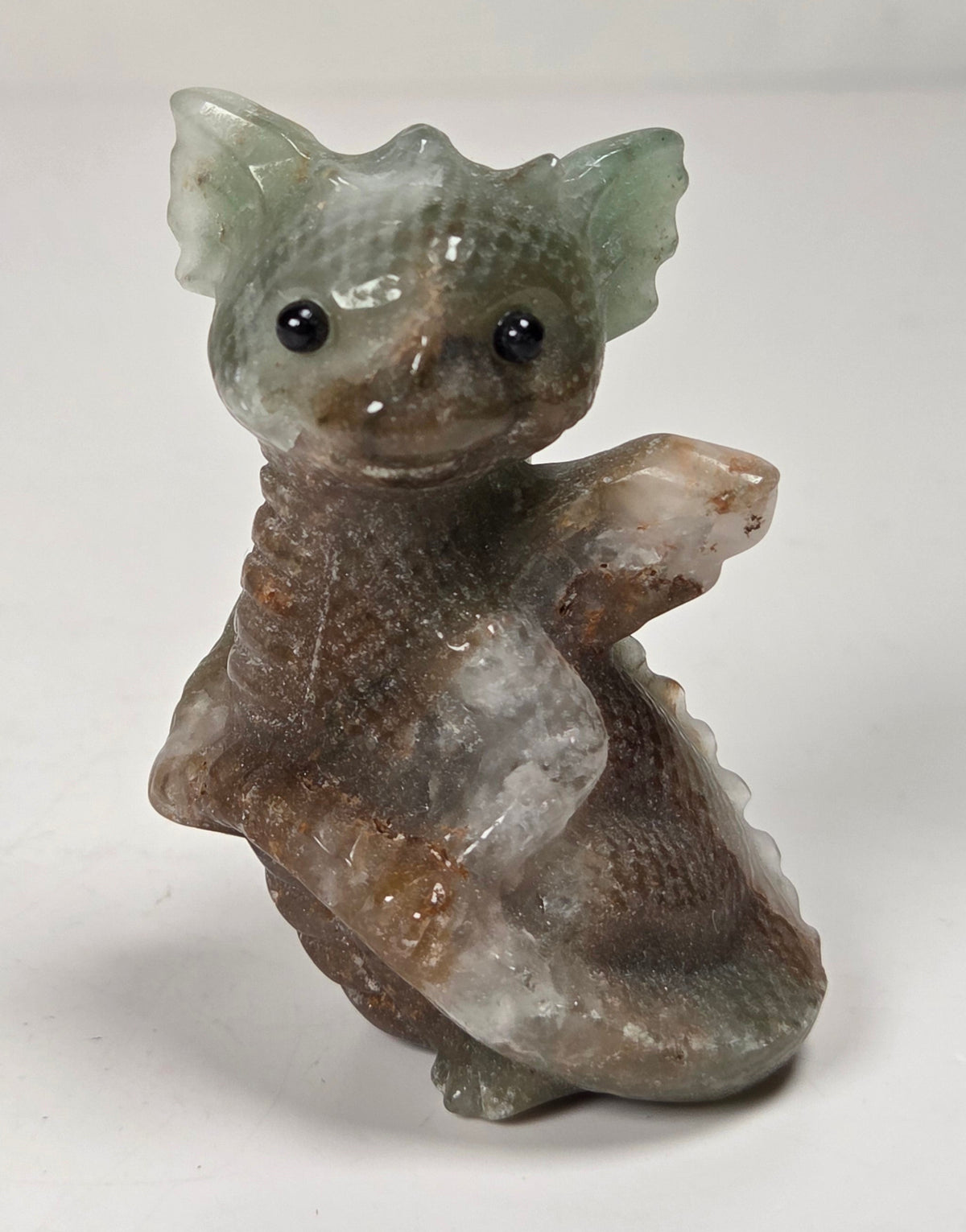 A small carved figurine of a creature with large ears, black eyes, and a textured, earthy body in shades of brown, gray, and pale green, standing on a white surface.