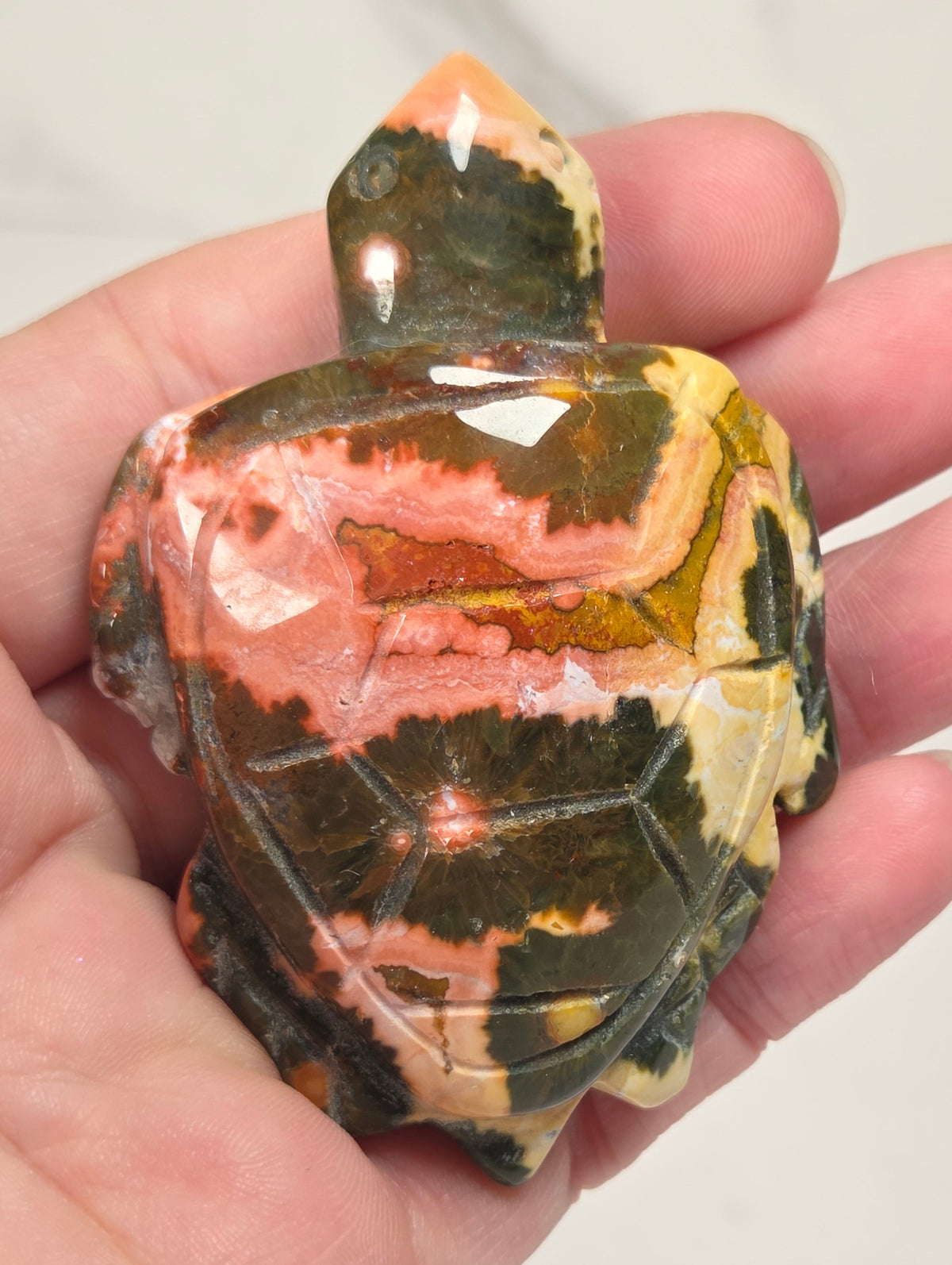 A hand holding a polished carved stone turtle figurine with marbled green, orange, pink, and cream patterns.