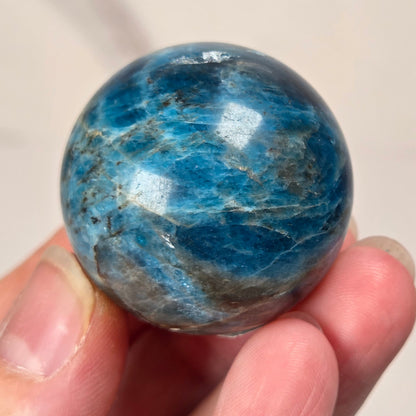 A polished blue gemstone sphere with natural marbling, being held in a hand, against a light marble background.