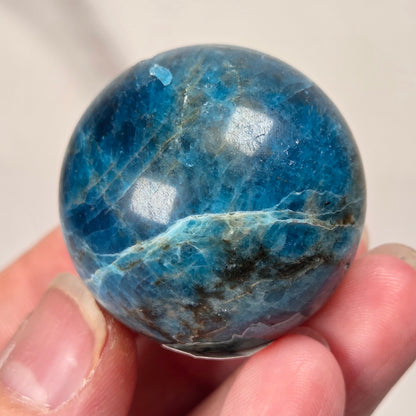 A polished blue gemstone sphere with natural marbling, being held in a hand, against a light marble background.