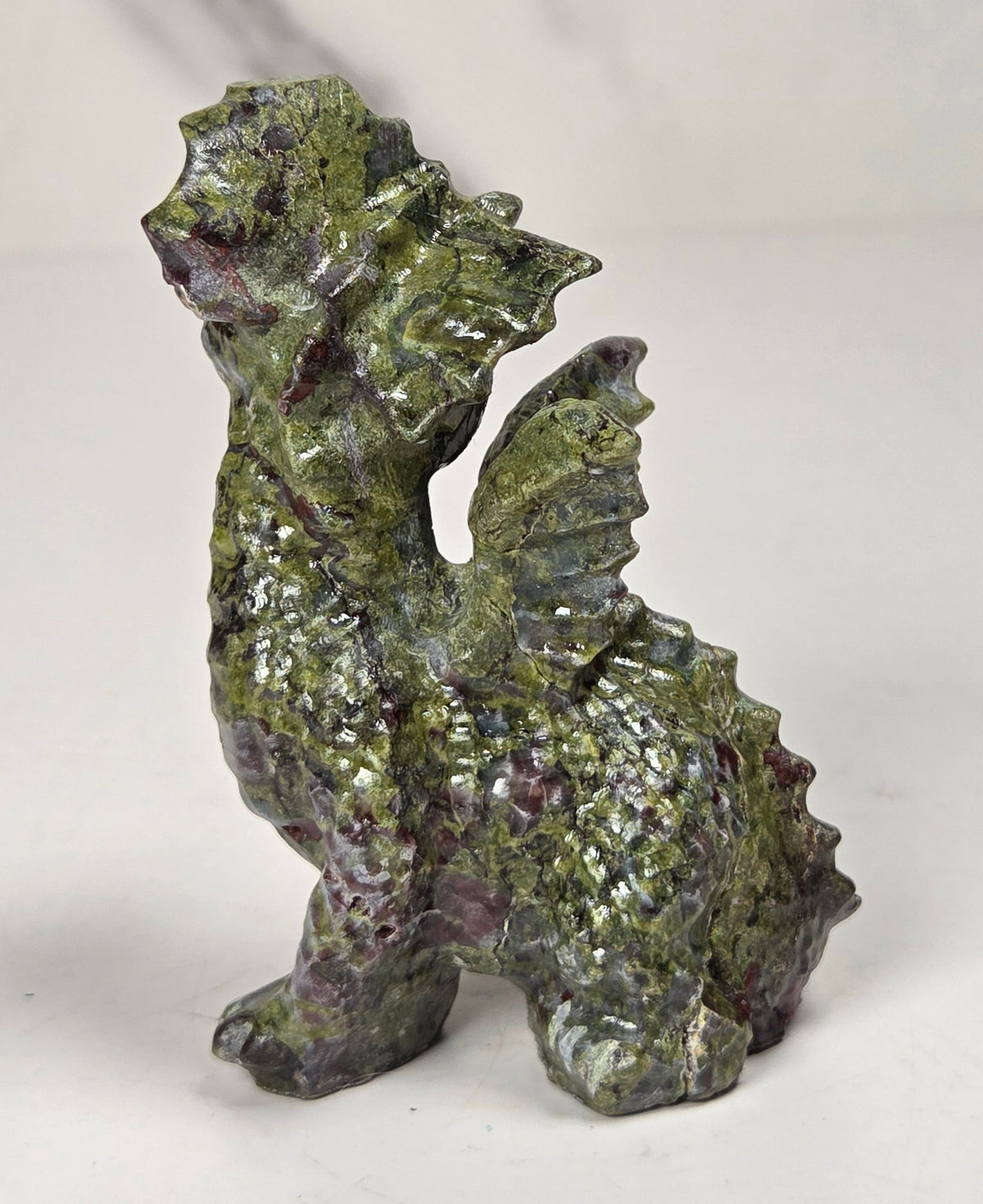 A small dragon figurine made from polished green and purple jasper stone, standing on a light gray surface with a white marble-patterned background.