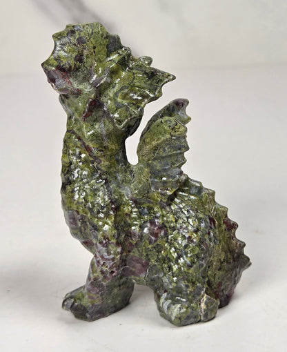 A small dragon figurine made from polished green and purple jasper stone, standing on a light gray surface with a white marble-patterned background.
