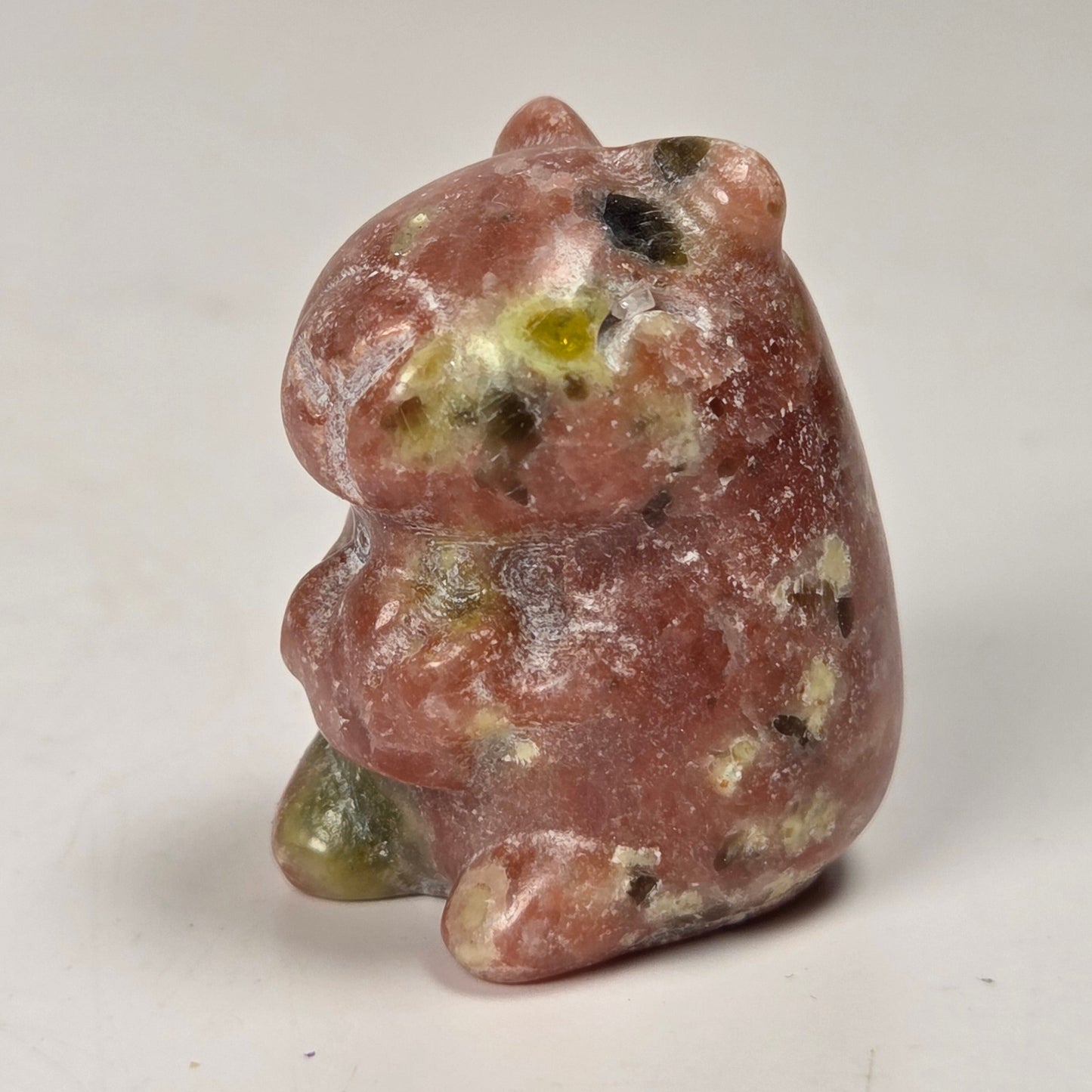 A small carved stone figurine of a bear, made from pinkish-red stone with green and white inclusions, set against a plain off-white background.
