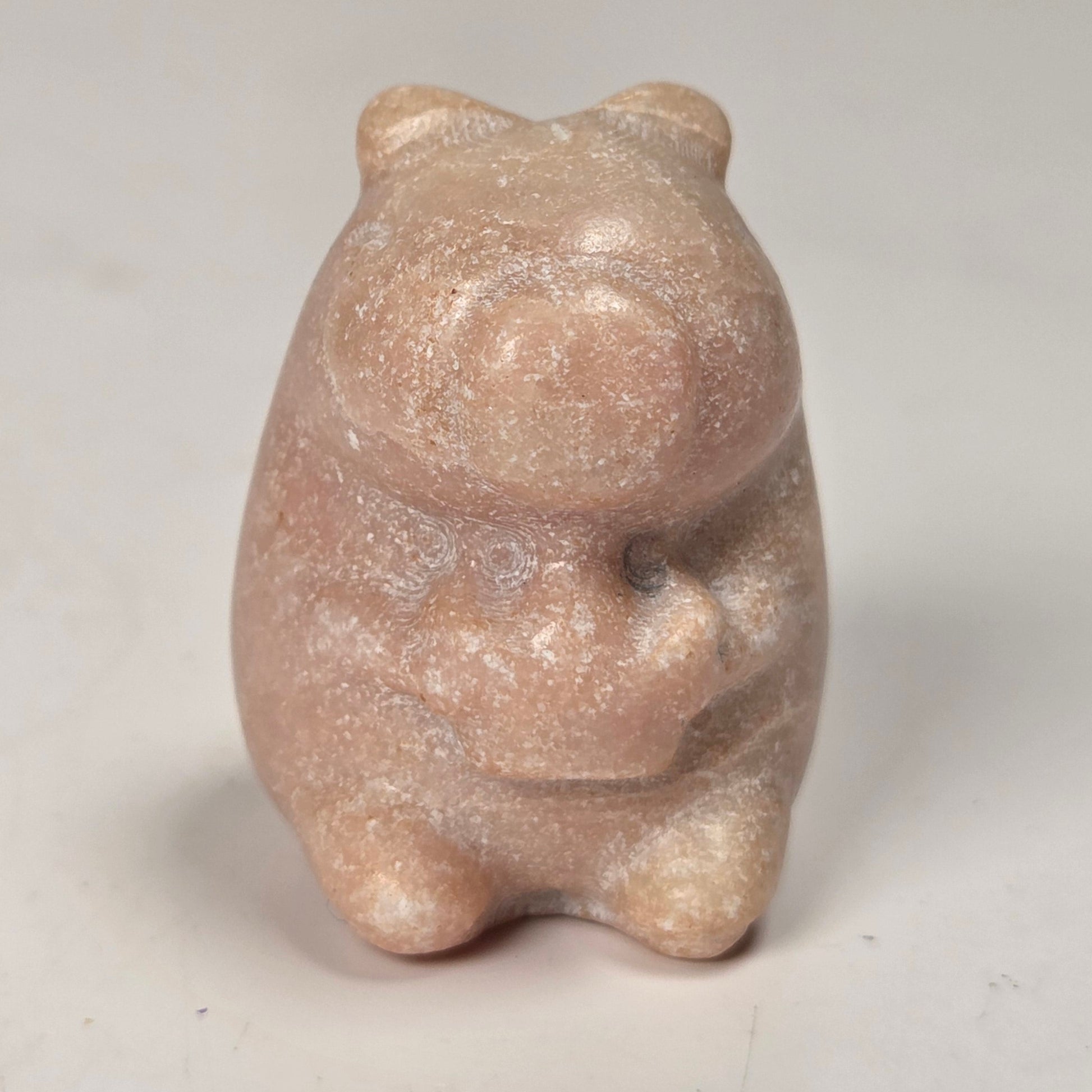 A small carved figurine of a bear or pig-like animal in a matte, dusty pinkish-beige stone or resin material, standing upright against a plain off-white background.