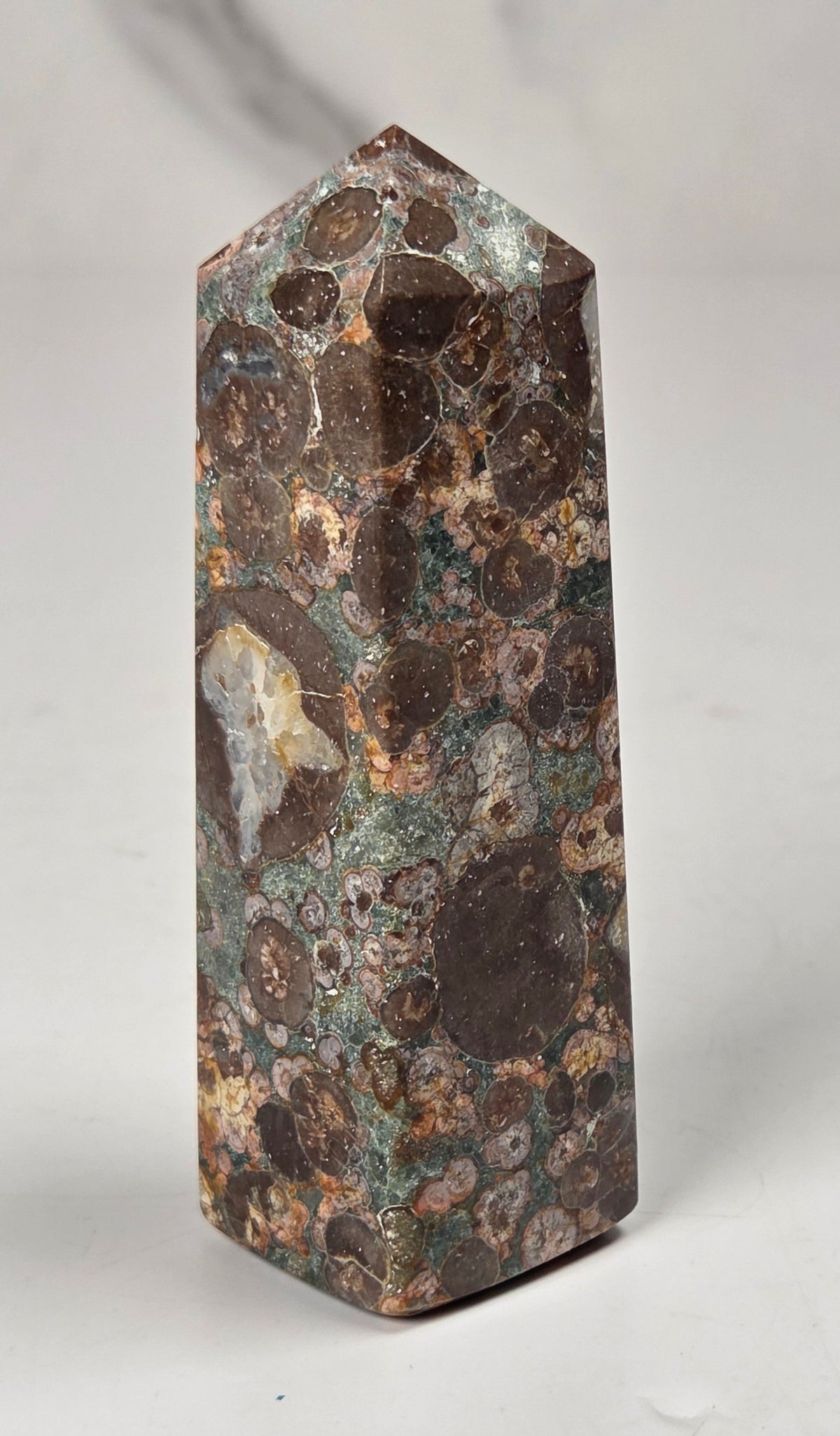 A polished natural mineral or stone tower with a pointed top, featuring a mottled pattern of dark brown, copper, and greenish textures, standing upright on a light gray surface.