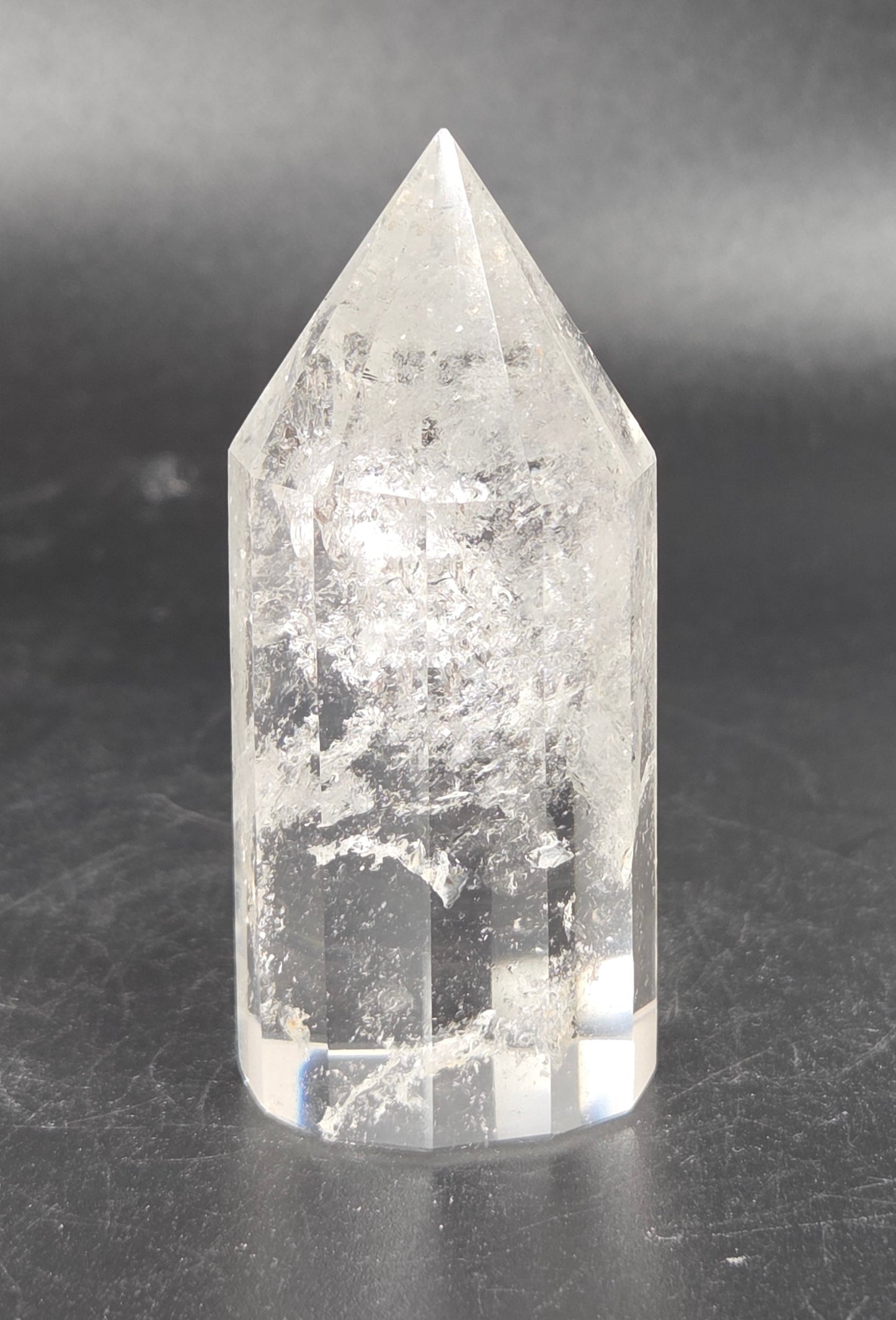 A clear quartz crystal point tower with natural inclusions, standing on a dark gray surface against a dark gray background.