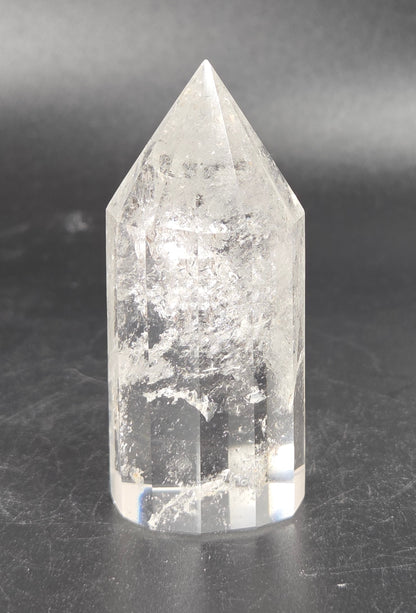A clear quartz crystal point tower with natural inclusions, standing on a dark gray surface against a dark gray background.