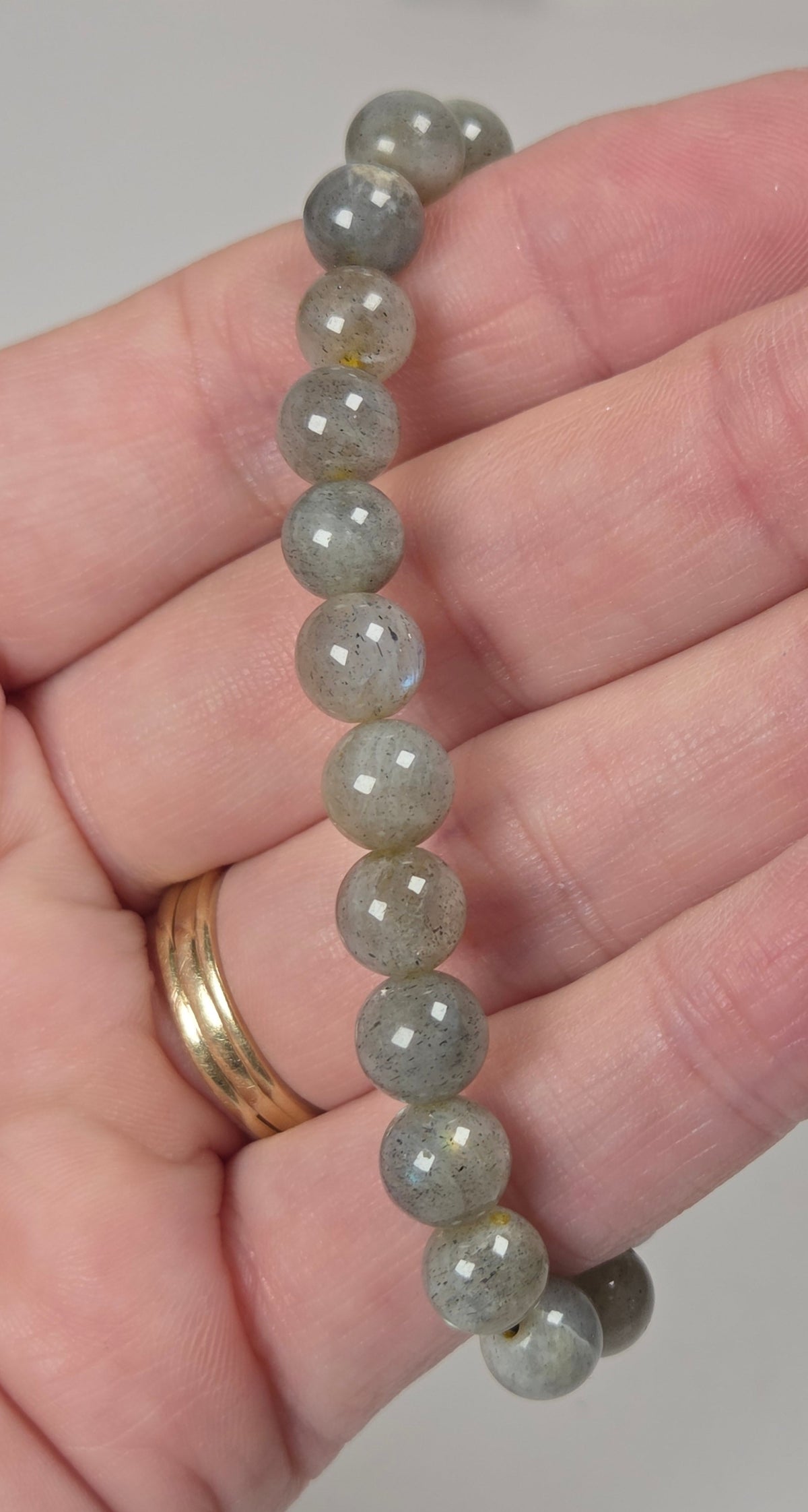 A beaded bracelet made of smooth, polished gray-green stone beads, shown resting on a person's hand. The hand also wears a gold ring.