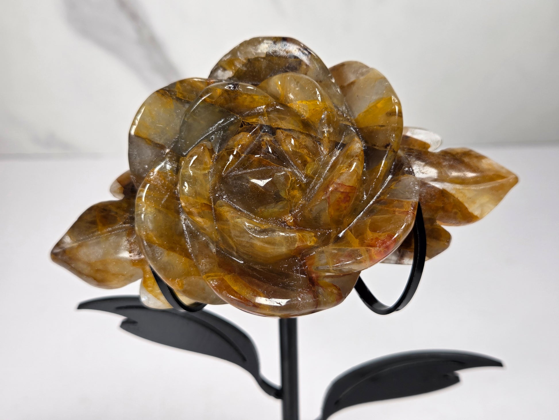 Golden Healer Crystal rose carving set on a black metal stand with leaves against a white background