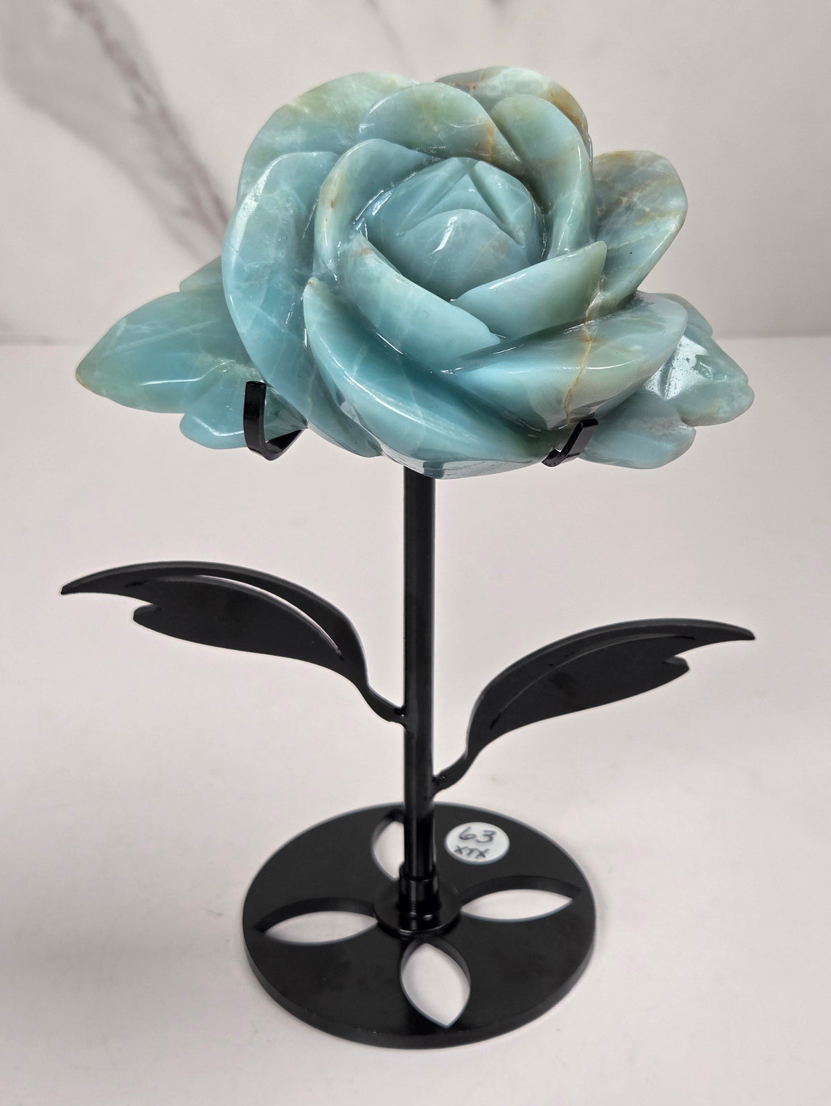 A carved light blue stone rose sculpture mounted on a black metal stand with leaf-like side supports and a round base, photographed against a light marble-patterned wall and white surface.