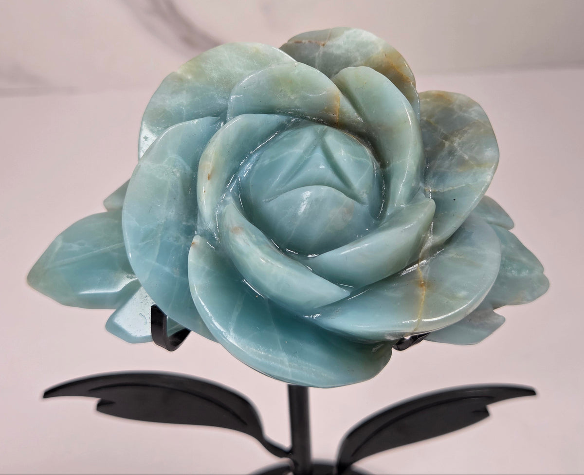 A carved light blue stone rose sculpture mounted on a black metal stand with leaf-like side supports and a round base, photographed against a light marble-patterned wall and white surface.