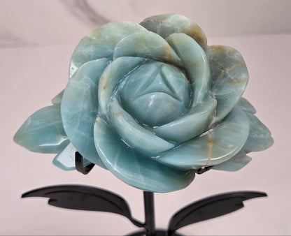A carved light blue stone rose sculpture mounted on a black metal stand with leaf-like side supports and a round base, photographed against a light marble-patterned wall and white surface.