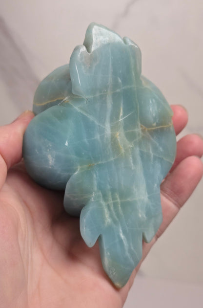 A carved light blue stone rose sculpture mounted on a black metal stand with leaf-like side supports and a round base, photographed against a light marble-patterned wall and white surface.