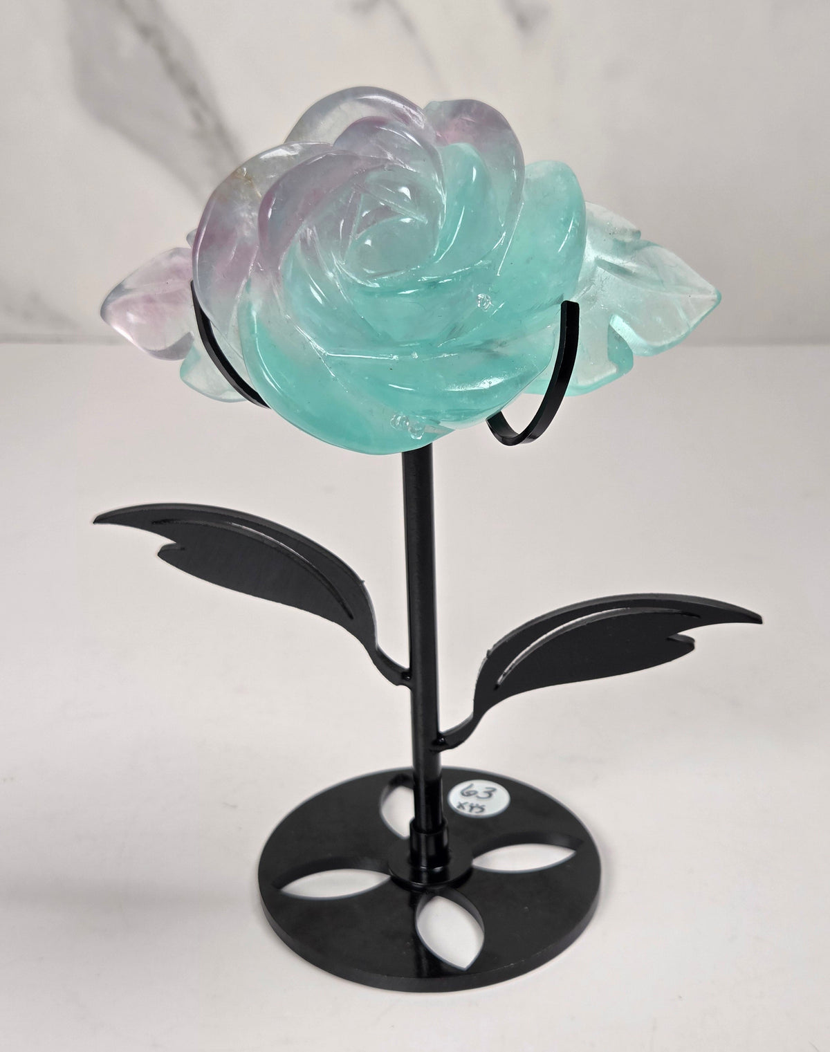 A decorative glass rose sculpture with iridescent pastel petals that shift between mint green and lavender, mounted on a black metal stem with leaves and a round base, photographed against a light marble-patterned background.