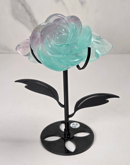 A decorative glass rose sculpture with iridescent pastel petals that shift between mint green and lavender, mounted on a black metal stem with leaves and a round base, photographed against a light marble-patterned background.