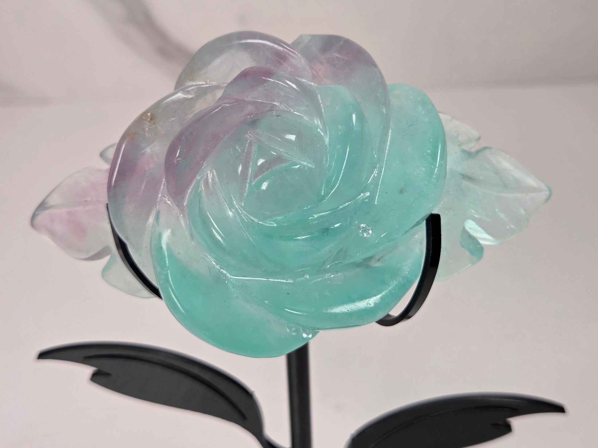 A carved rose-shaped decorative object made of translucent stone with a color gradient from light aqua blue to soft lavender-pink, mounted on a black metal stand against a plain light background.