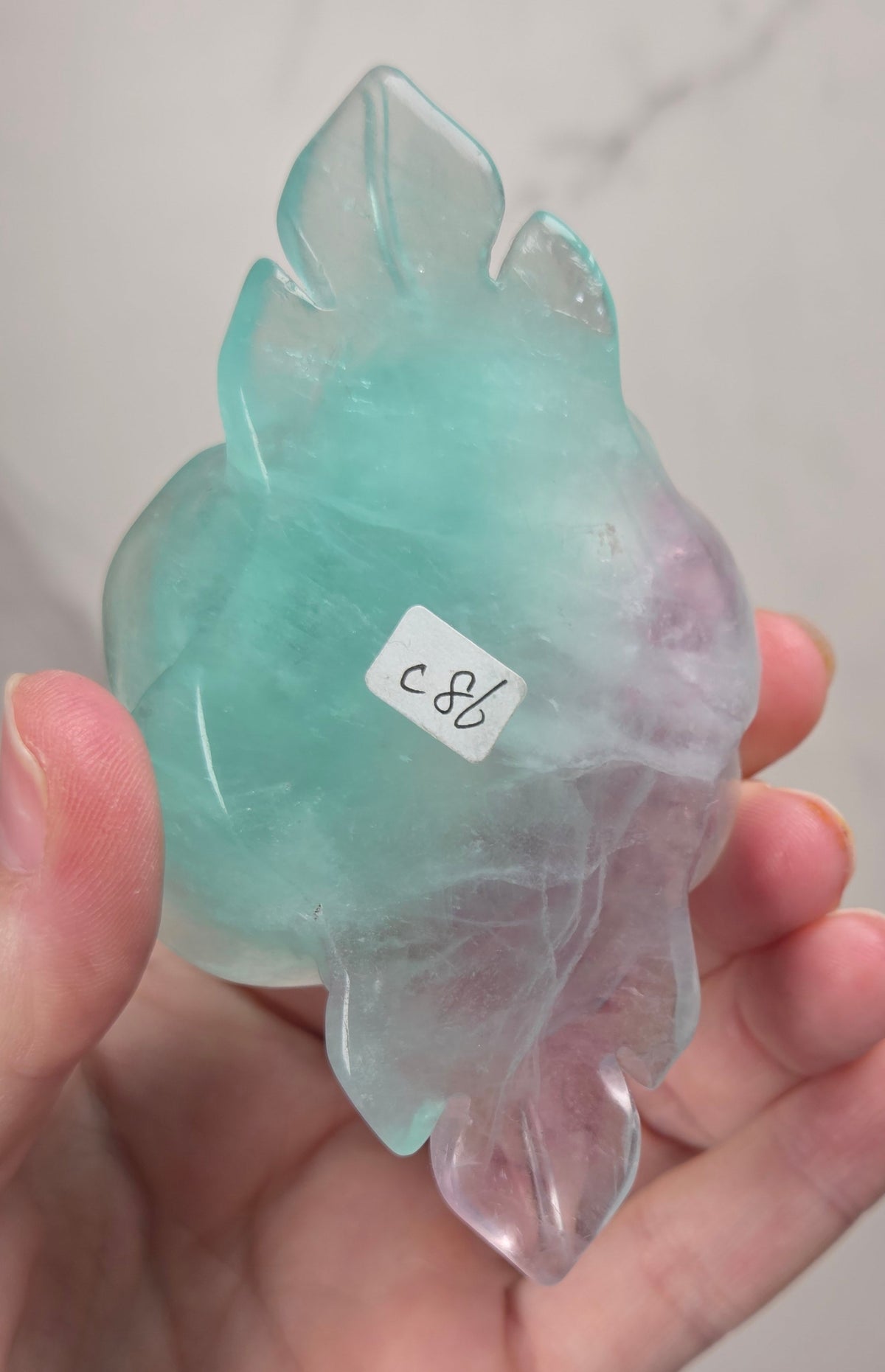 A hand holds a carved crystal or stone figurine with a leaf-like shape. The stone is translucent with a light aqua/turquoise center fading into pale pink and lavender at the edges.