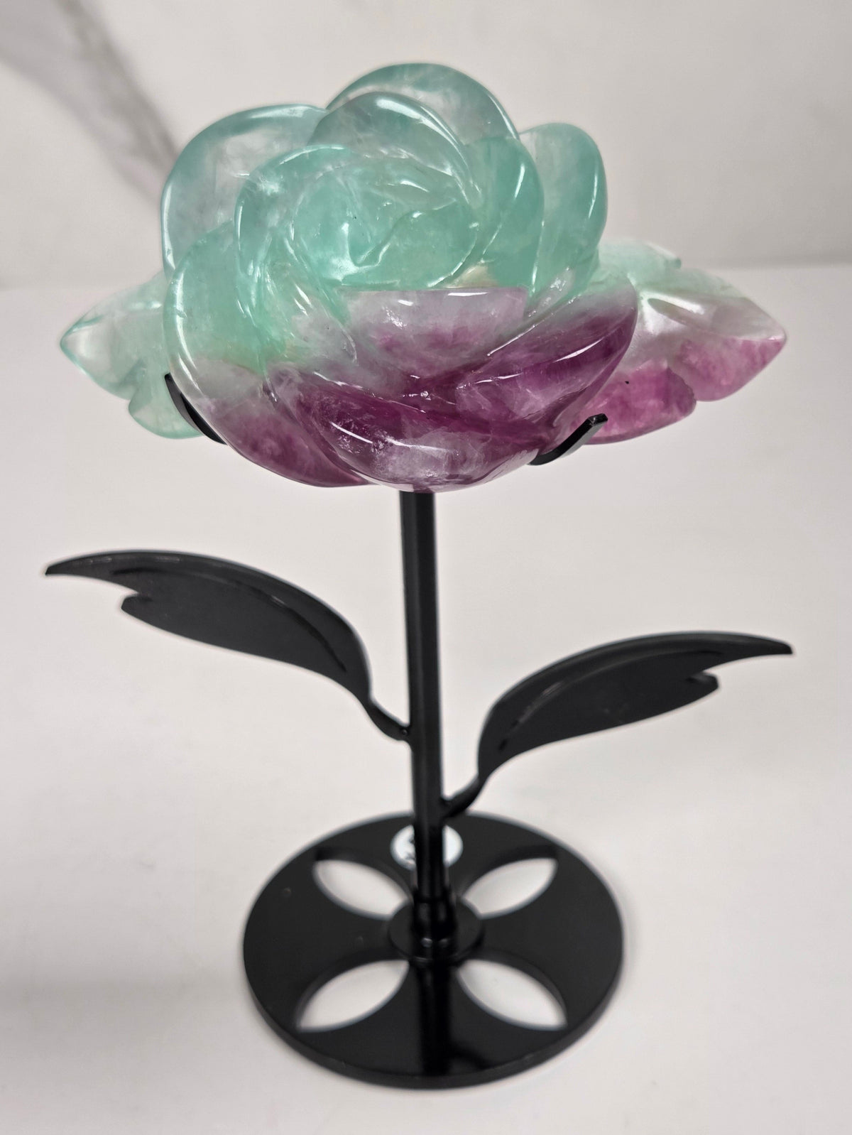A carved rose-shaped decorative object made of translucent stone with a color gradient from light aqua blue to soft lavender-pink, mounted on a black metal stand against a plain light background.