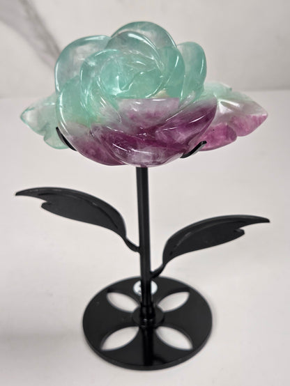A carved rose-shaped decorative object made of translucent stone with a color gradient from light aqua blue to soft lavender-pink, mounted on a black metal stand against a plain light background.
