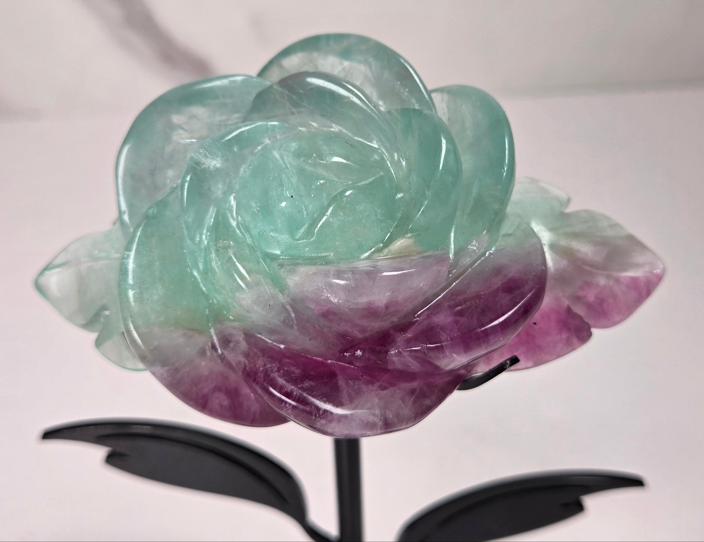 A carved rose-shaped decorative object made of translucent stone with a color gradient from light aqua blue to soft lavender-pink, mounted on a black metal stand against a plain light background.