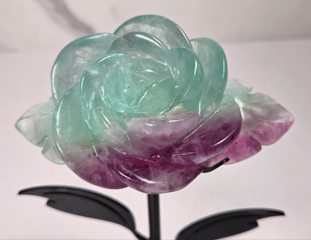 A carved rose-shaped decorative object made of translucent stone with a color gradient from light aqua blue to soft lavender-pink, mounted on a black metal stand against a plain light background.