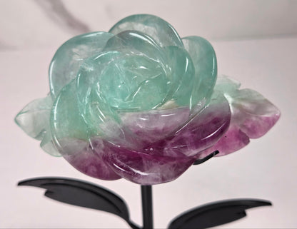 A carved rose-shaped decorative object made of translucent stone with a color gradient from light aqua blue to soft lavender-pink, mounted on a black metal stand against a plain light background.