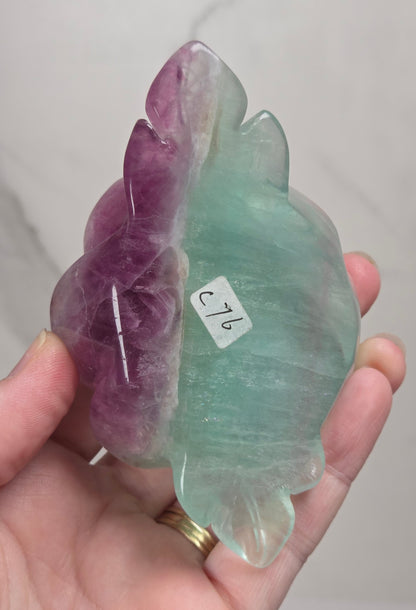 A carved rose-shaped decorative object made of translucent stone with a color gradient from light aqua blue to soft lavender-pink, mounted on a black metal stand against a plain light background.