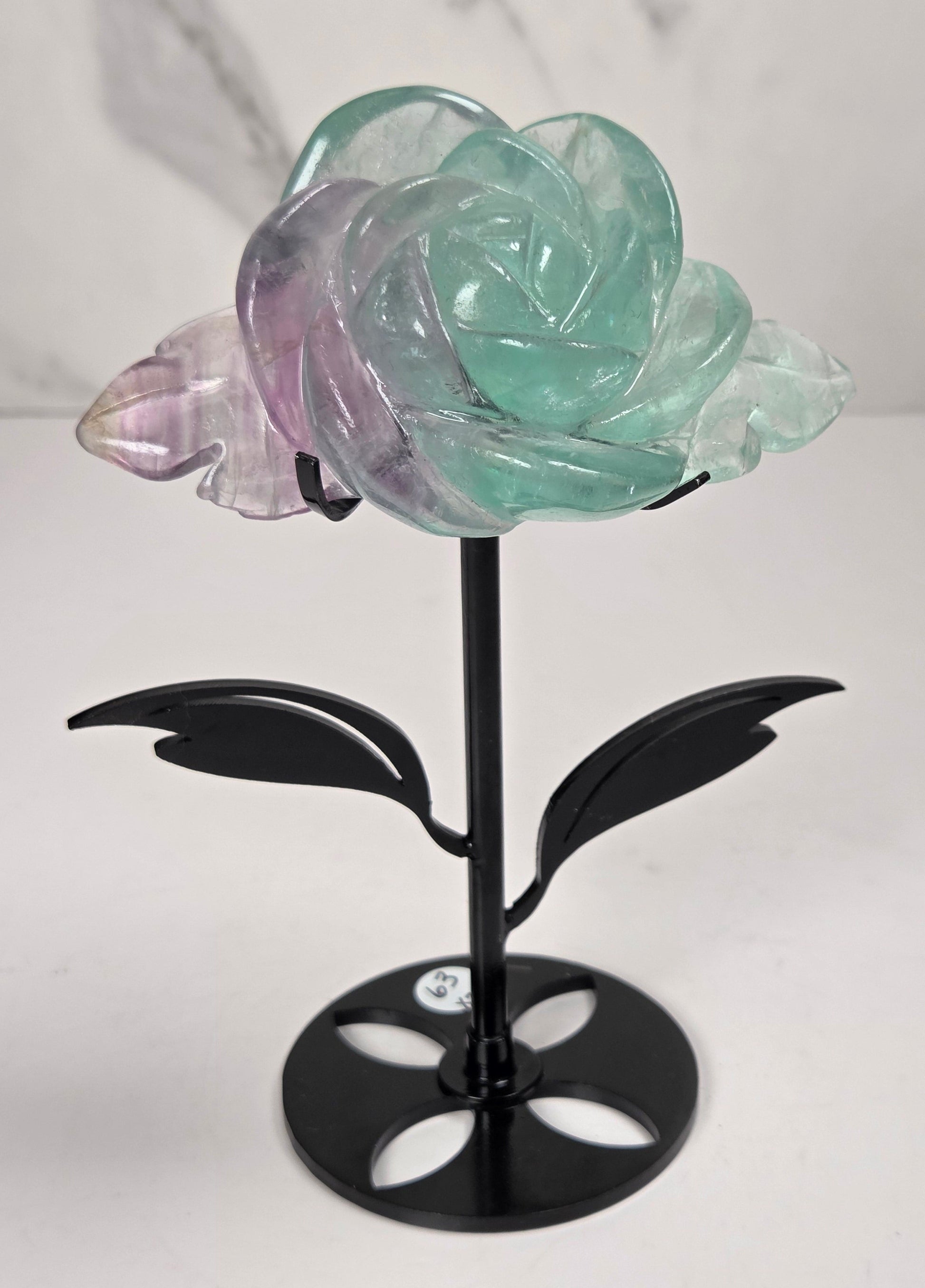 A carved rose-shaped decorative object made of translucent stone with a color gradient from light aqua blue to soft lavender-pink, mounted on a black metal stand against a plain light background.