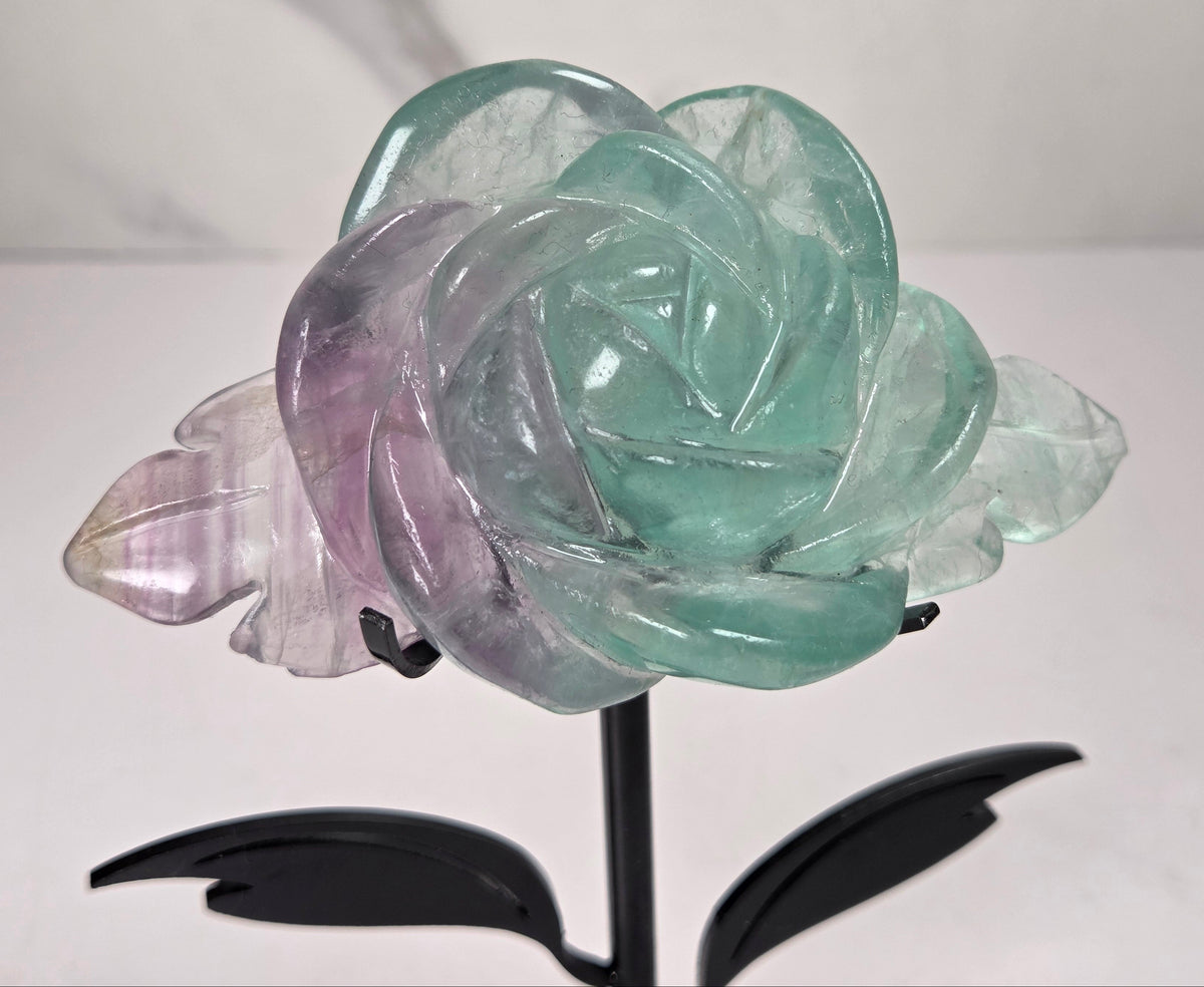 A carved crystal rose sculpture with layered petals in mint green, lavender purple, and pale pink, mounted on a black metal stand with two leaves.
