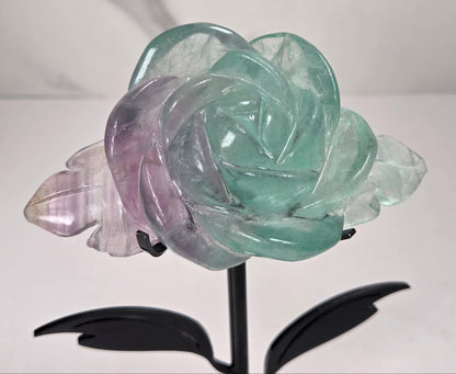 A carved crystal rose sculpture with layered petals in mint green, lavender purple, and pale pink, mounted on a black metal stand with two leaves.