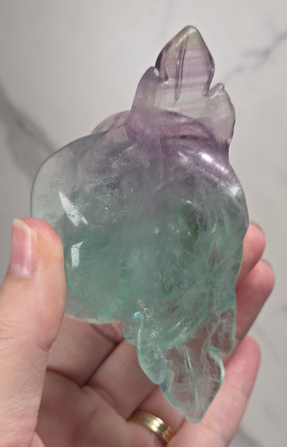 A carved rose-shaped decorative object made of translucent stone with a color gradient from light aqua blue to soft lavender-pink, mounted on a black metal stand against a plain light background.