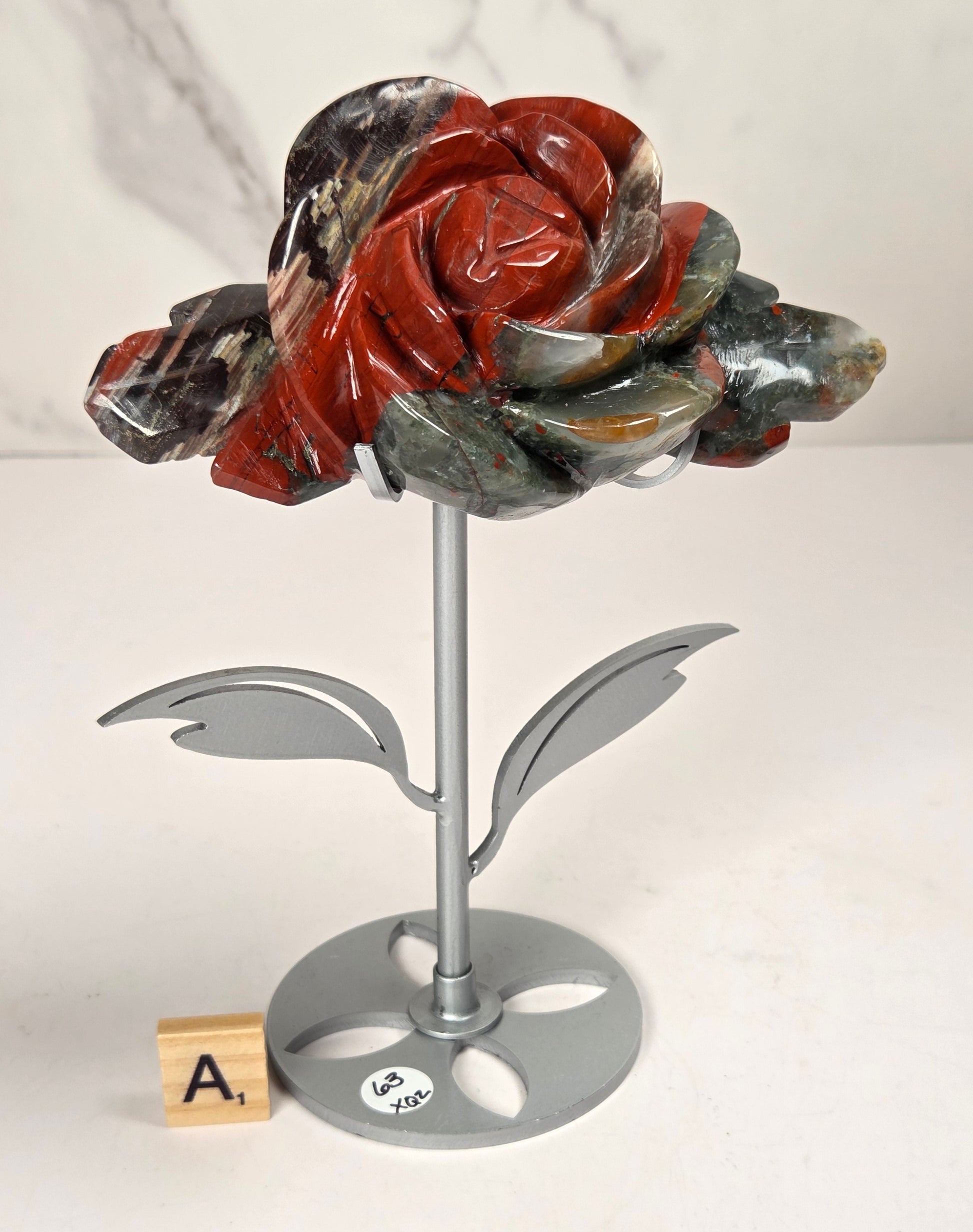 A carved stone rose sculpture with red, brown, and gray marbled petals on a silver metal stem and round base, displayed on a white surface with a small wooden letter tile nearby.