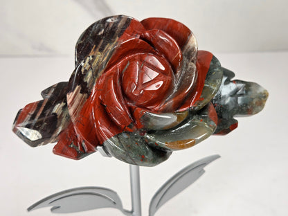 A carved stone rose sculpture with red, brown, and gray marbled petals on a silver metal stem and round base, displayed on a white surface with a small wooden letter tile nearby.