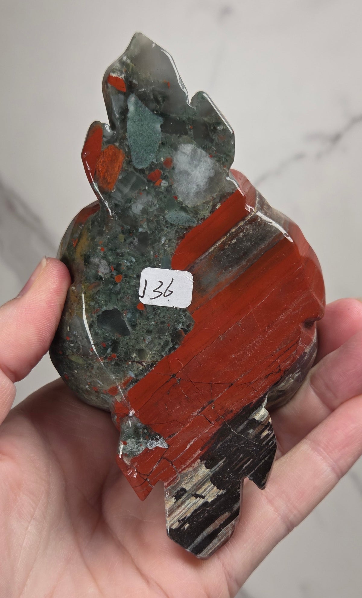 A hand holds a polished natural stone specimen with layered red, green, gray, and black patterns, featuring a small white sticker with the number 136 on it, set against a light marble background.